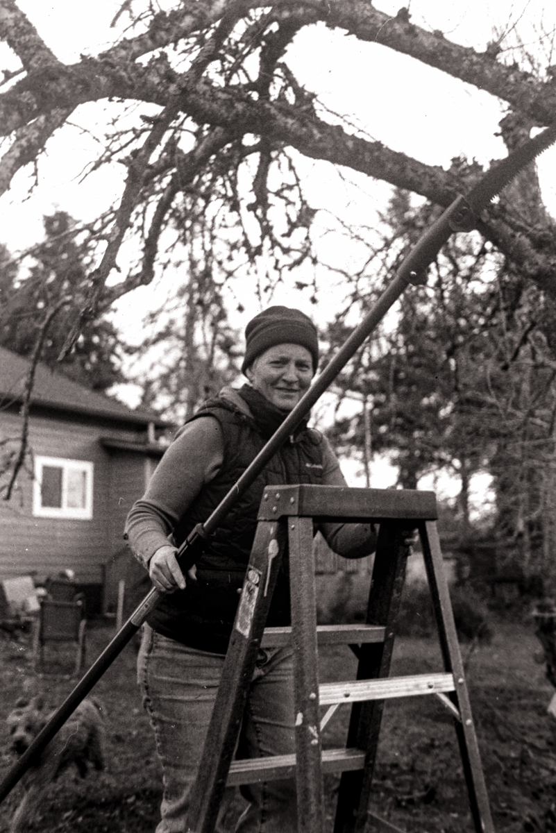 A black and white film image of suzie pruning an apple tree and another looking up at some poplar trees.